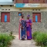 Two mature women standing by one another and dressed in typical Congolese women fashion in the from of CEEDECO stone-made building.